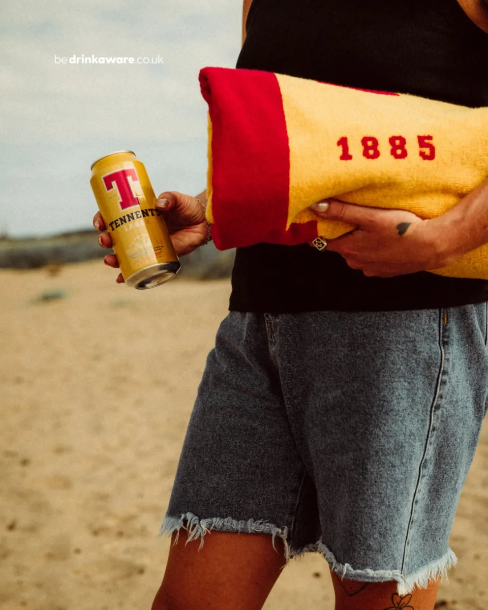 Person holding a can of Tennent's Lager and a towel on a beach.